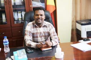 A man in a plaid shirt is sitting at a wooden desk with papers, a water bottle, a calendar, and a sanitizer bottle. Behind him are shelves with files, an Ethiopian flag, and a printer on a cabinet. He is smiling and holding a pen.