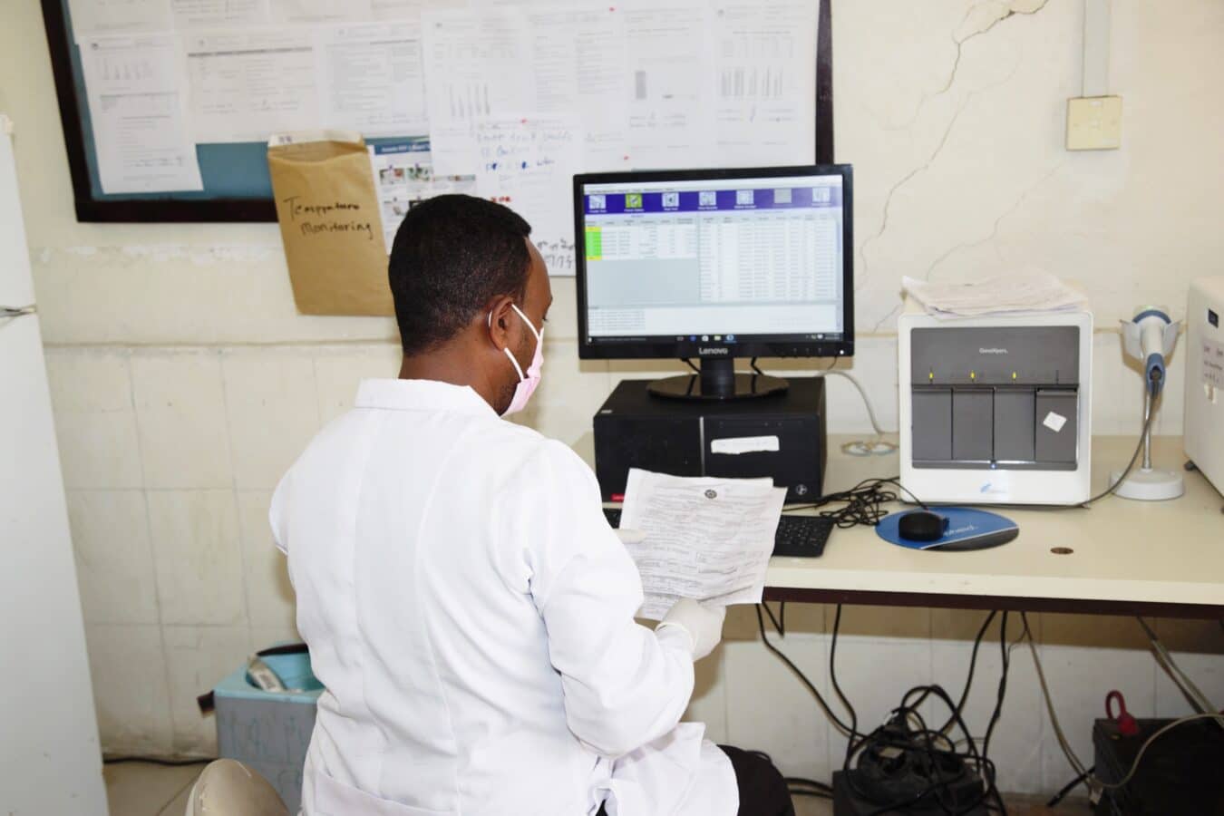 A person wearing a white lab coat and a face mask sits at a desk, analyzing data on a computer screen. The desk is cluttered with papers and a printer. The wall is decorated with pinned documents and charts.