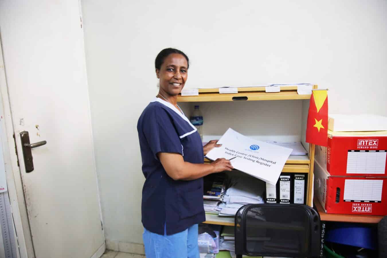 A nurse in a blue uniform smiles at the camera while organizing papers on a shelf in a tidy office space. A small flag and stacked boxes are visible on the shelf.