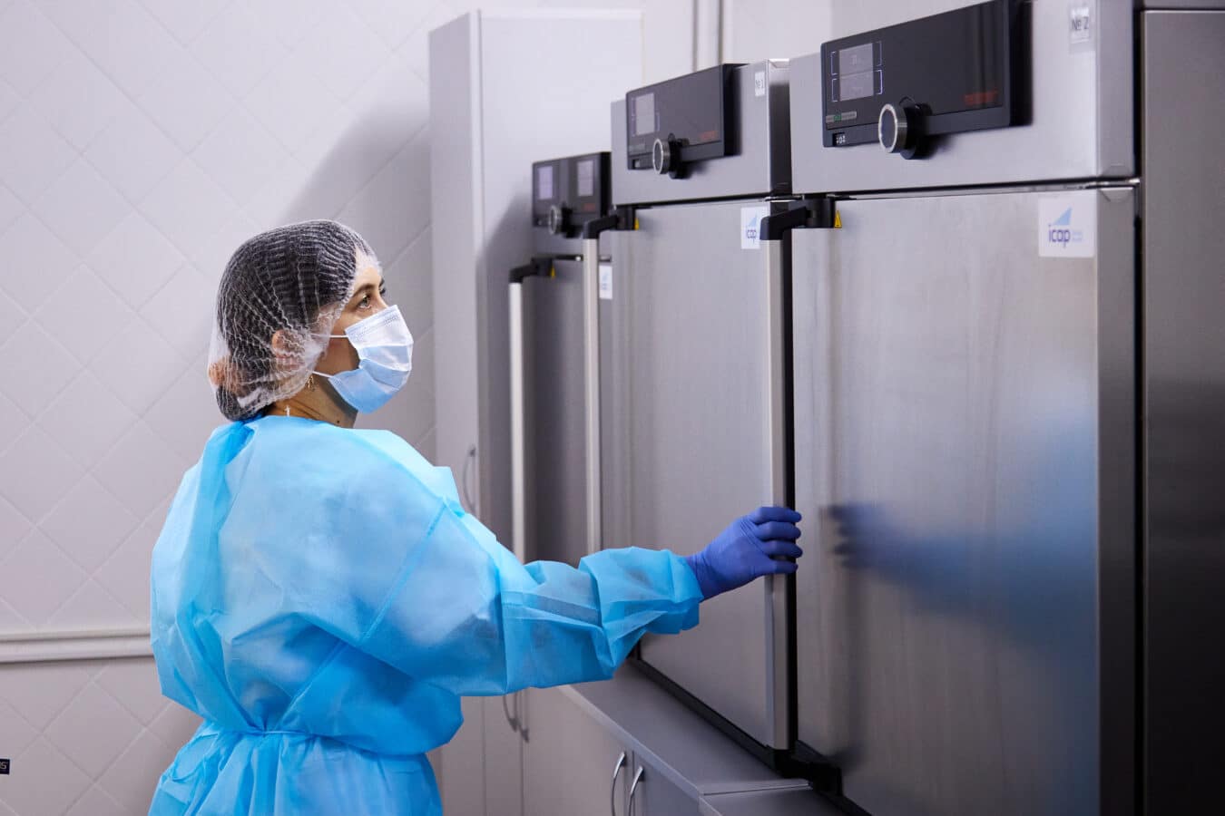 A person wearing a blue protective gown, hair net, face mask, and gloves opens a stainless steel laboratory incubator or storage unit. The room has white tiled walls and additional similar equipment.