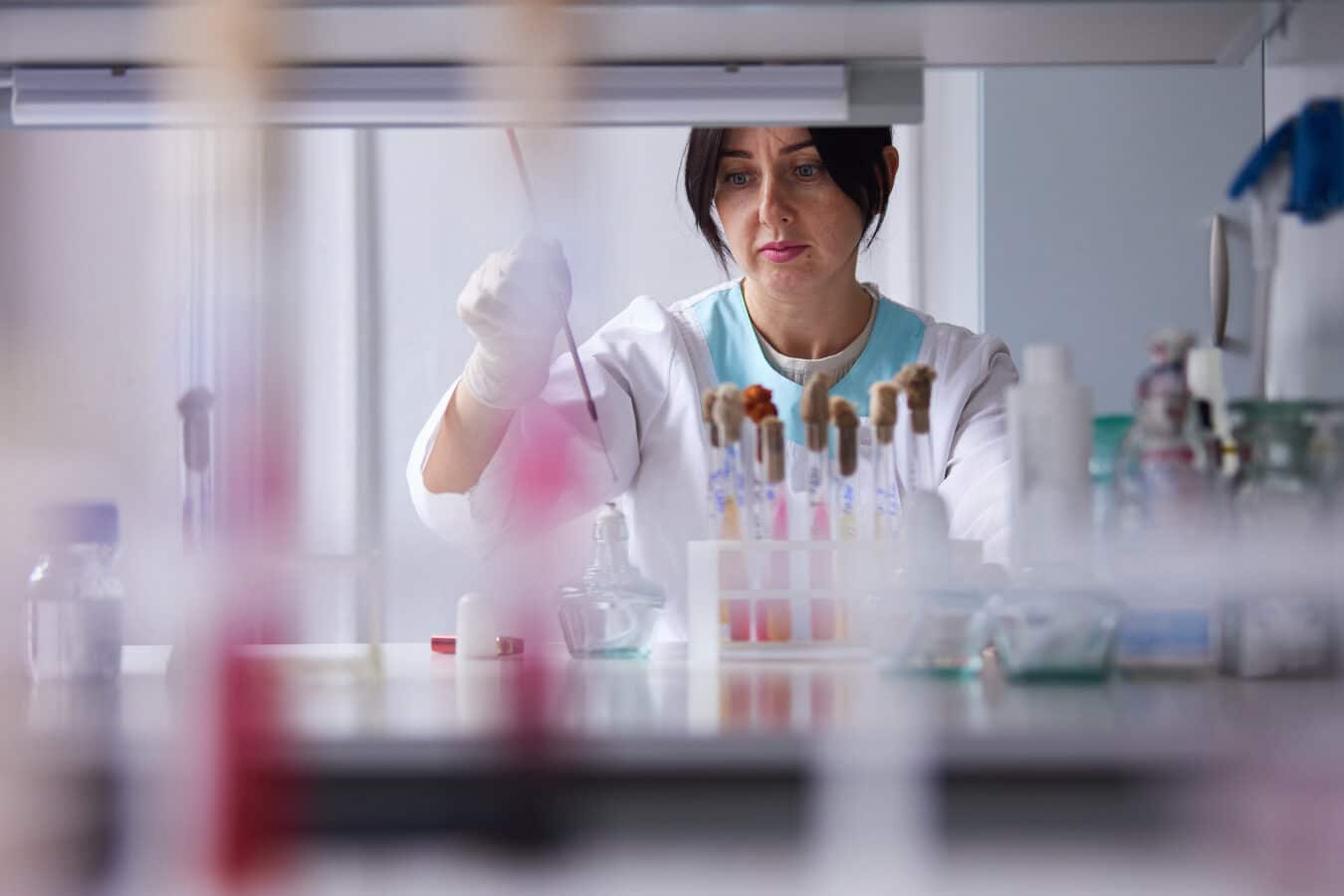 A scientist wearing a white coat and gloves works in a laboratory, holding a pipette over test tubes filled with colorful liquids on a bench. The background is blurred, showing various lab equipment.