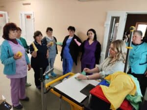 A group of women, some in uniforms, attentively watch a woman seated at a cleaning cart as she demonstrates how to use colored cloths for cleaning. The women each hold a colored cloth.