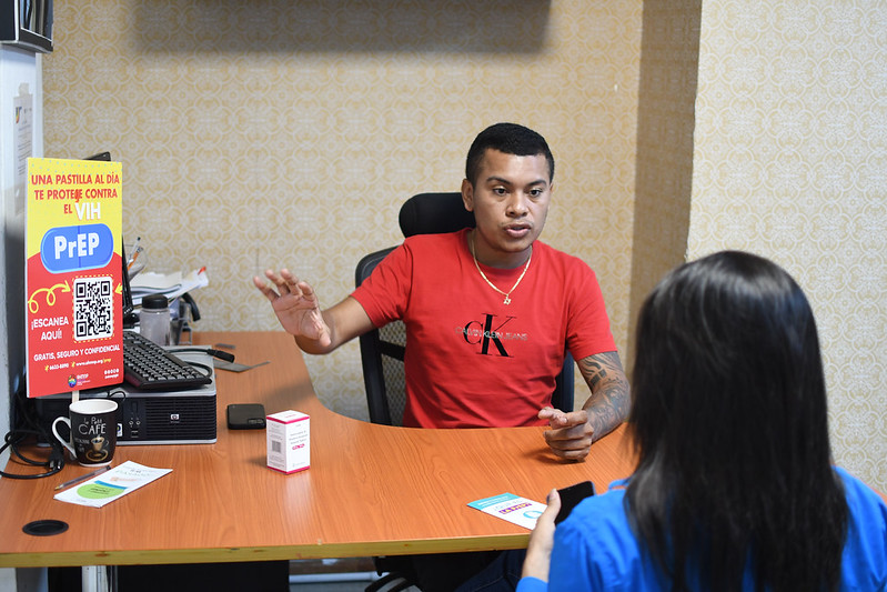 A man in a red shirt sits at a desk talking to a woman with dark hair facing him. On the desk is a sign with information about PrEP.