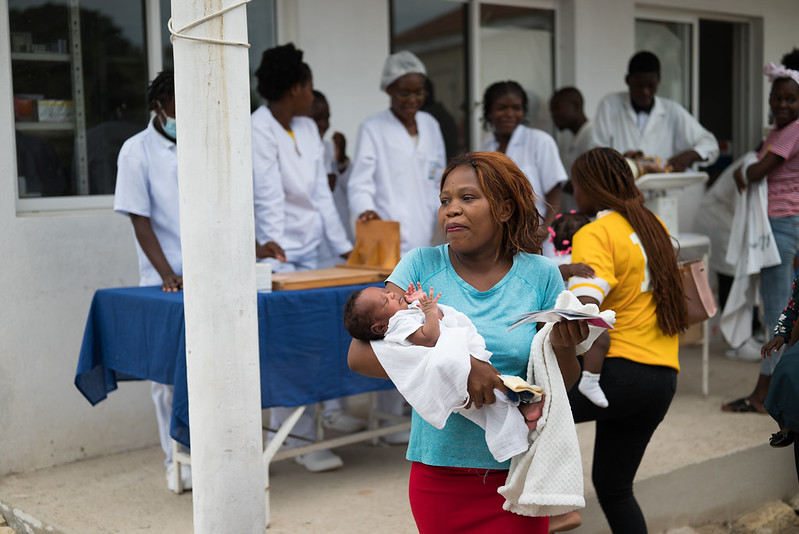 A woman holds a baby in front of table of health care workers in white outfits