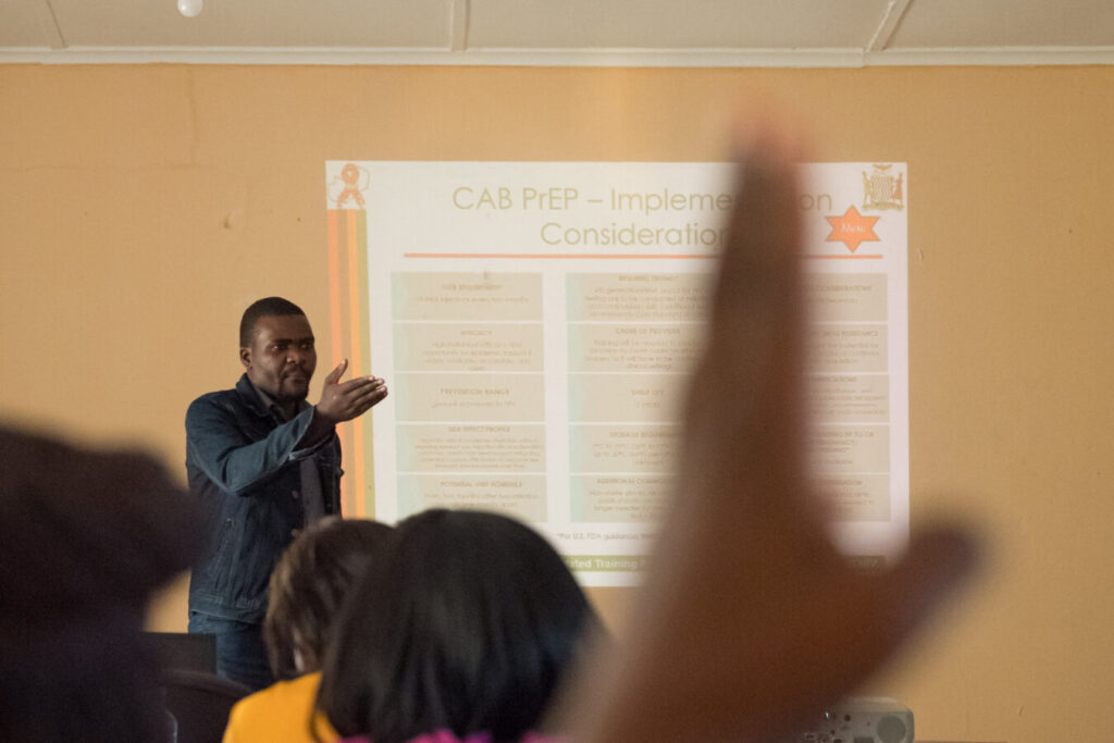 A man lectures to a room; a hand is raised in the foreground