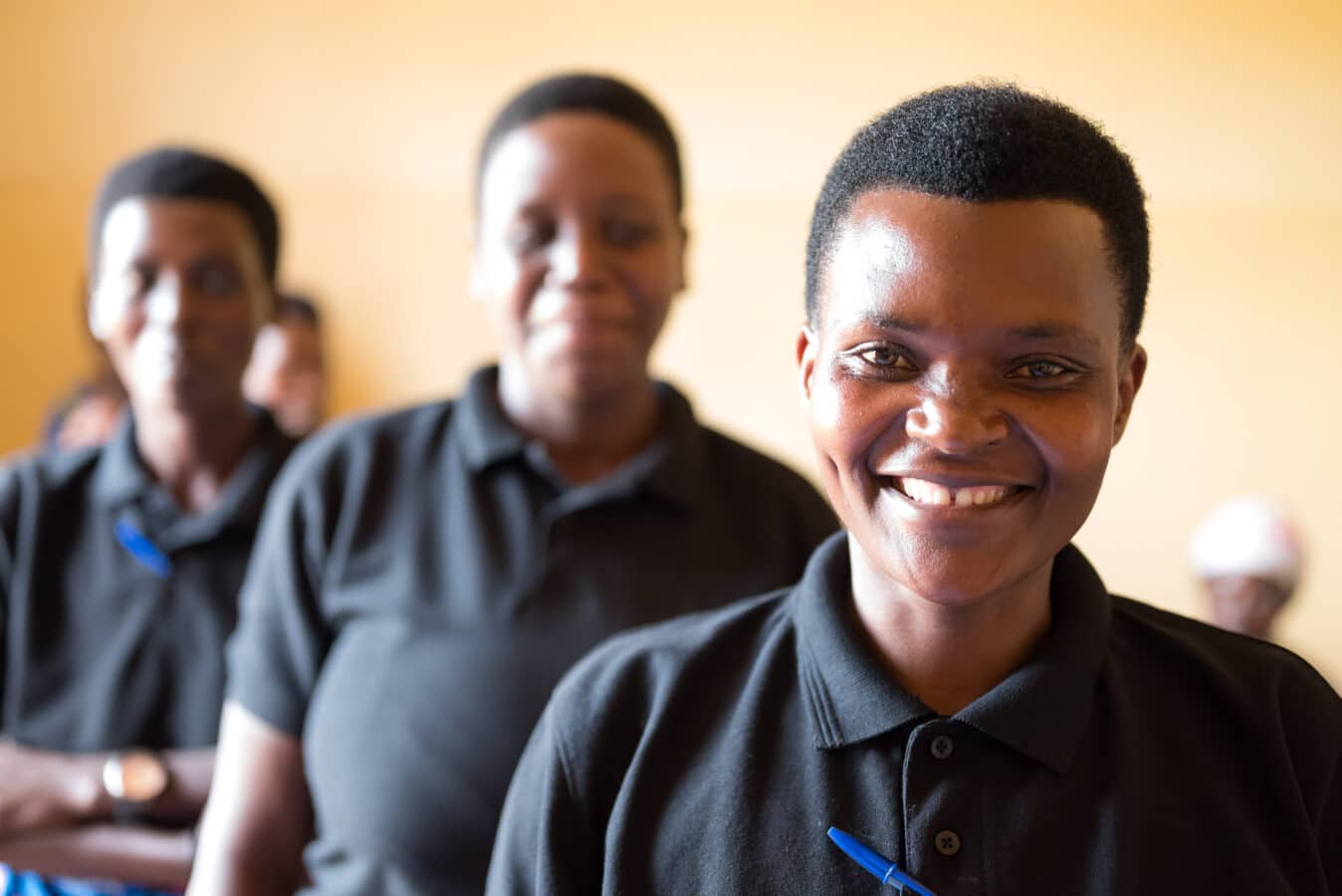 Four people are standing in a line, all wearing black polo shirts. The person at the front, who is smiling, is in focus, while the others are slightly blurred in the background. The setting appears to be indoors with a light-colored wall.