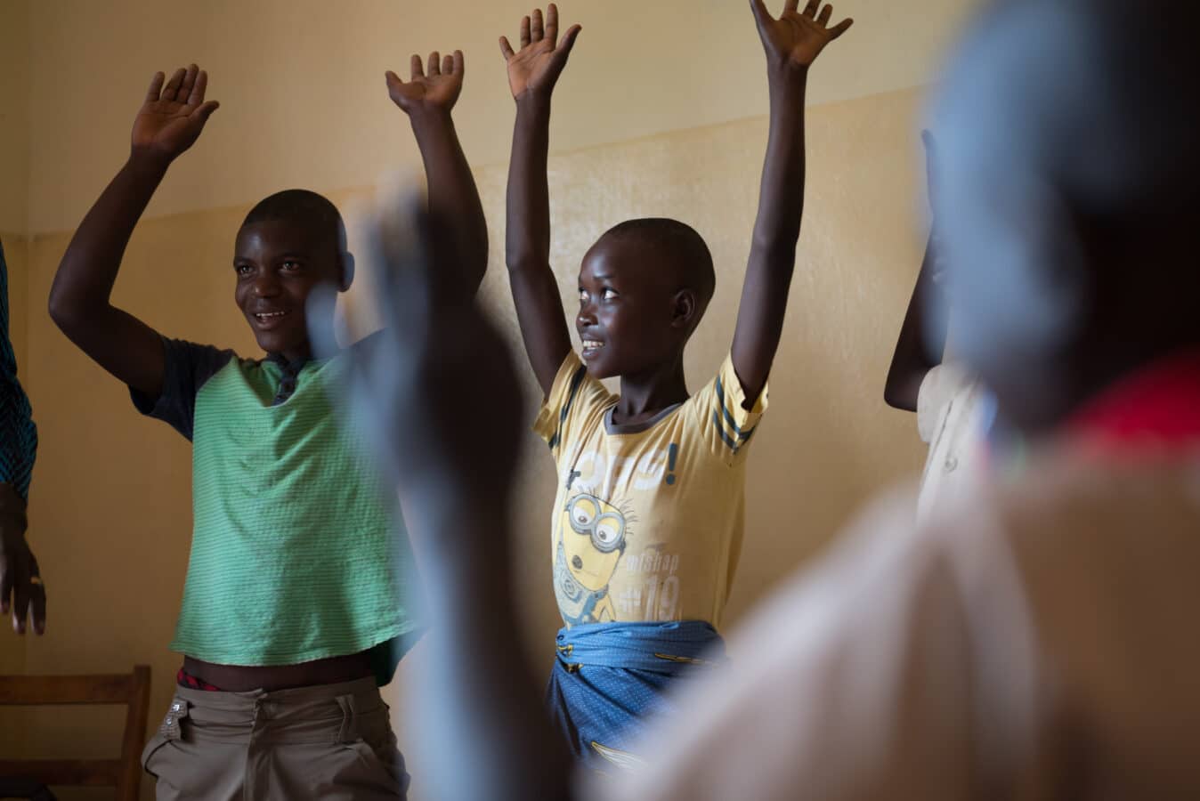 A group of children stand indoors with their hands raised above their heads, smiling and engaged in activity. The room has light-colored walls, and the children appear joyful and enthusiastic.