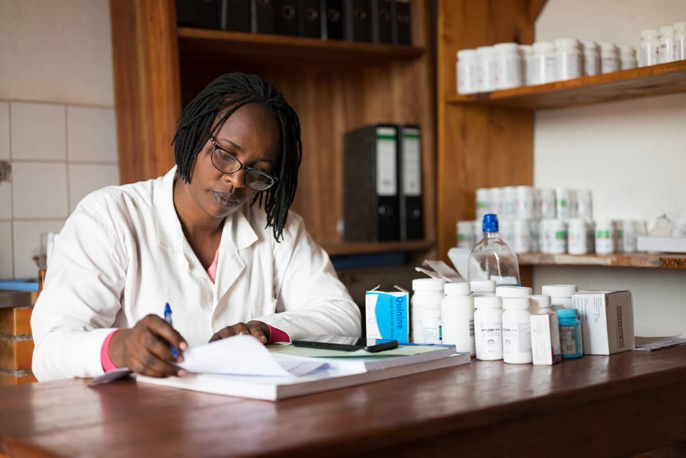 A pharmacist wearing a white coat sits at a desk in a pharmacy. She is writing in a notebook surrounded by prescription medicine bottles and various drug packages on the desk. Shelves filled with more medication are in the background.