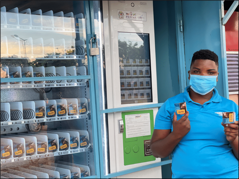 A person wearing a blue shirt and face mask stands next to a vending machine, holding two small boxes in their hands. The vending machine has multiple rows of slots filled with similar boxes.