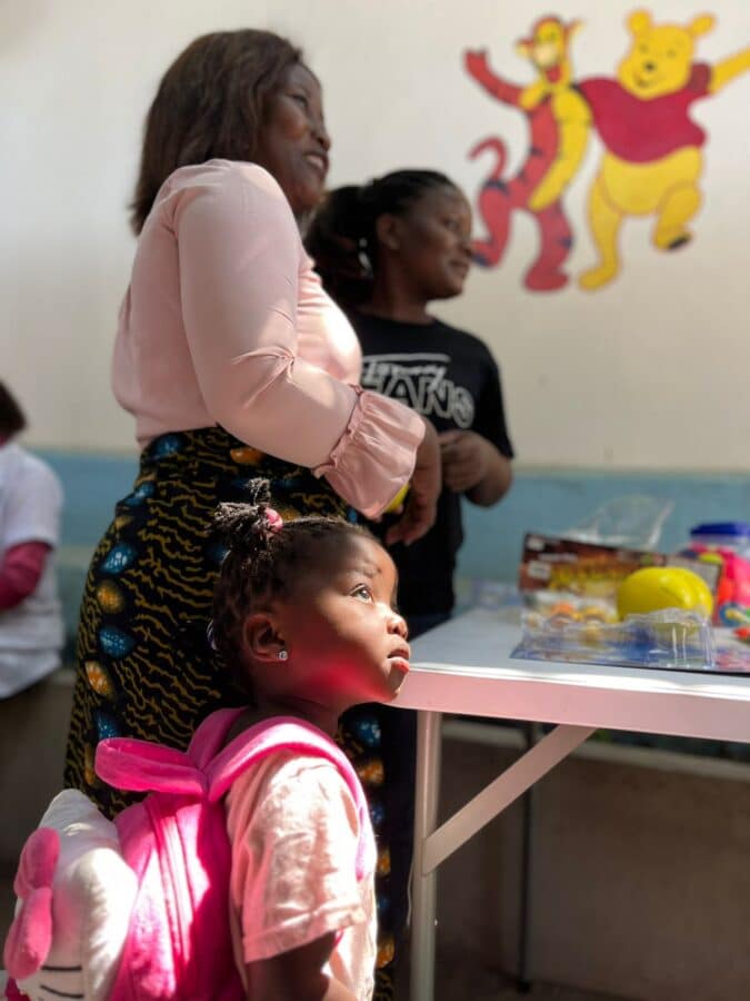 A young girl with a pink backpack looks up while standing next to a woman in a bright room. Another person is in the background near a table with items on it. The wall features a colorful mural of Winnie the Pooh and Tigger.