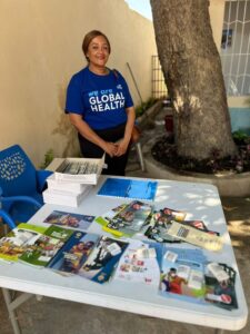 A woman stands behind a table covered with pamphlets and brochures about healthcare. She is wearing a blue shirt that reads "We are Global Health." A large tree and a building are in the background.