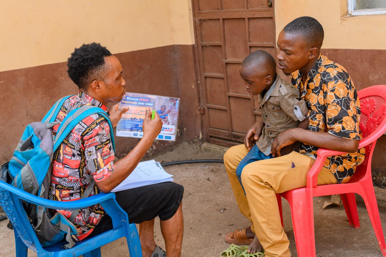 A community health worker sits on a blue chair, showing a pamphlet to a man and his young son seated on a red chair. They are in an outdoor setting with a brown door and beige wall in the background.