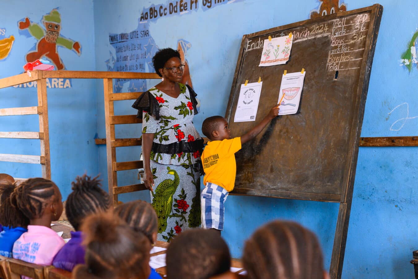 A teacher stands beside a young student pointing at a drawing on a blackboard in a classroom. Children are seated, watching attentively. The room is colorful, with educational posters and murals on the walls.