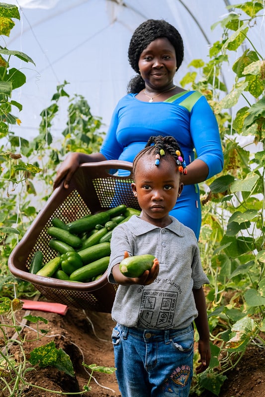 A woman in a blue dress stands in a greenhouse holding a basket of cucumbers. In front of her, a young girl with beaded braids offers a cucumber towards the camera. Green plants surround them.