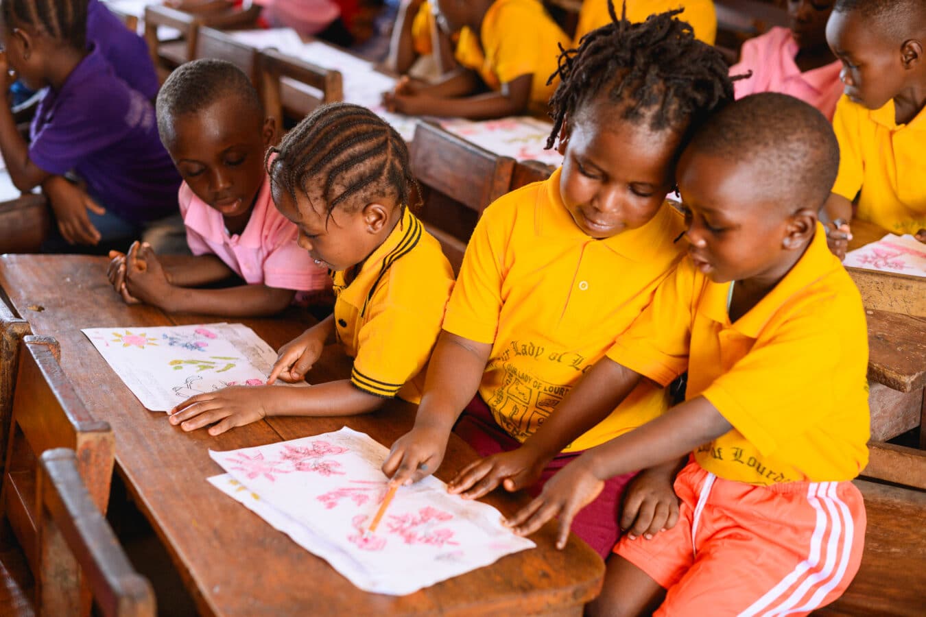 Four young children in school uniforms are seated at wooden desks, focused on coloring with crayons. They are in a brightly lit classroom, each holding a paper with floral designs. The atmosphere is lively and educational.