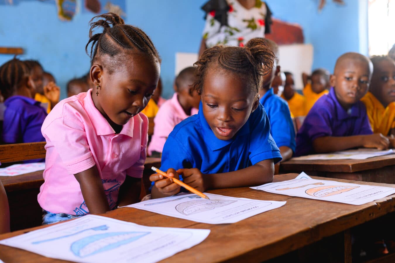 Two young girls in pink and blue shirts are smiling and working together at a wooden desk, surrounded by classmates in colorful uniforms. They are engaged in a learning activity, focusing on worksheets with outlines of teeth on them.