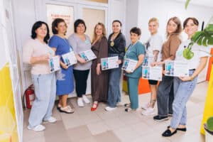 Eight smiling women stand indoors in a hallway, holding certificates and posing for a group photo. There is a door and a fire extinguisher in the background, and a large green plant to the right.