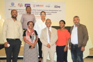 A group of seven people stands in front of a banner at an event. They are smiling and dressed in business casual attire. The banner behind them features logos and text related to a pilot implementation dissemination and national-scale-up workshop organized by health organizations.