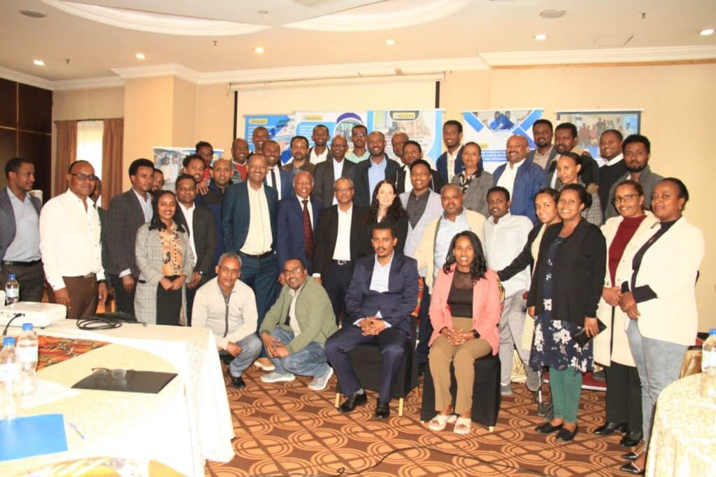 A group photo shows around 40 people gathered in a room. Everyone is dressed in business or smart casual attire. Some are sitting in the front row while others are standing. There are presentation banners in the background and tables in the foreground.