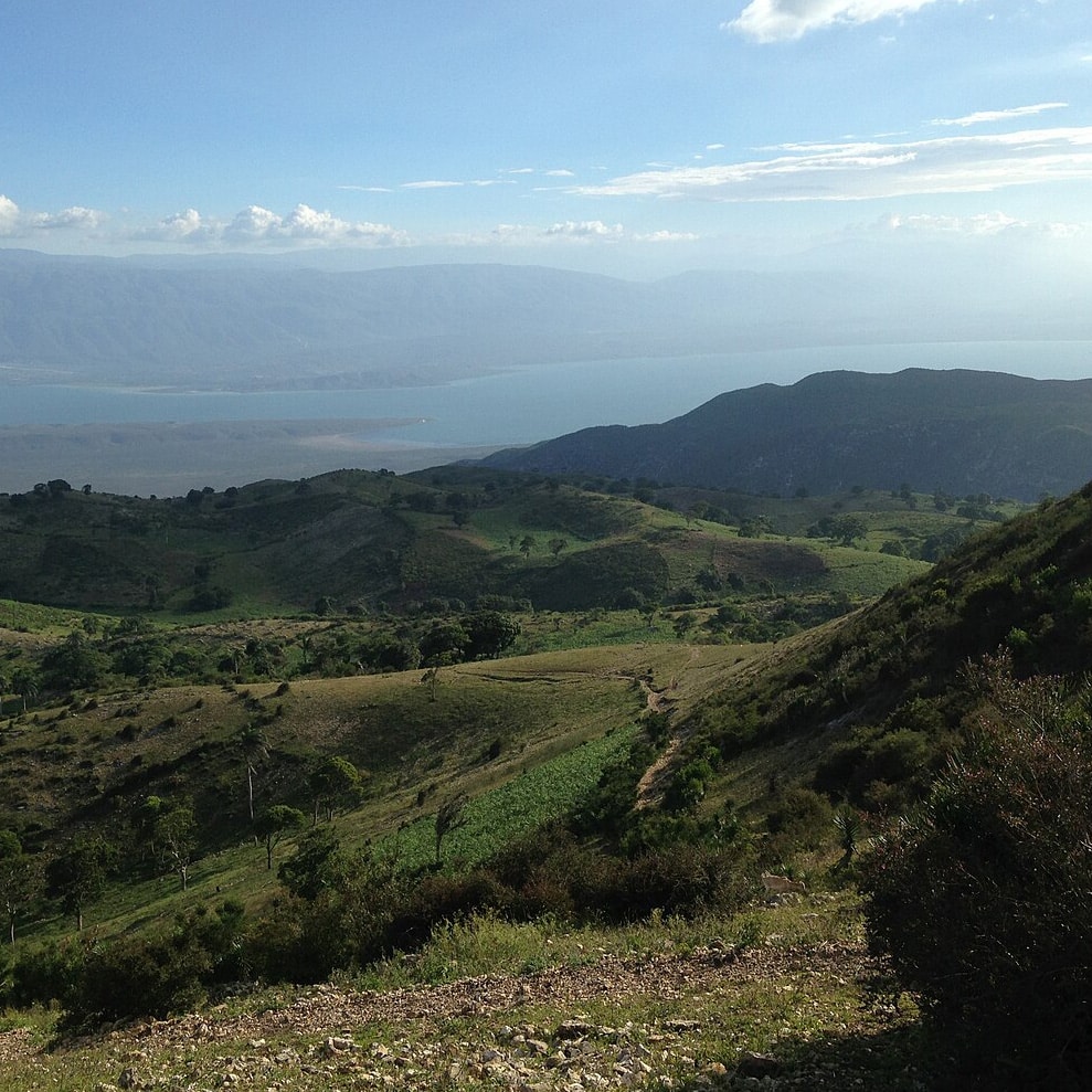 A scenic view of rolling green hills with sparse trees, leading down to a large body of water in the background. The sky is clear with a few scattered clouds, and distant mountains are visible on the horizon.