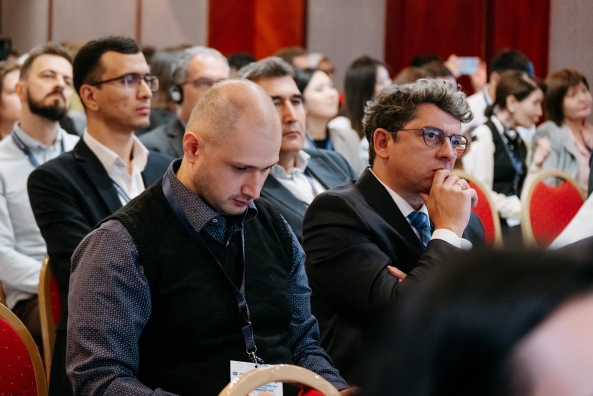 A group of people sitting attentively in a conference room on red chairs. The attendees, mostly men in professional attire, are focused on the speaker. One man in the front appears to be taking notes, while others listen intently.