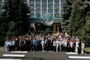 A large group of professionally dressed people pose for a photo outside a building with a glass entrance. The background includes green trees and the facade of the multi-story building.