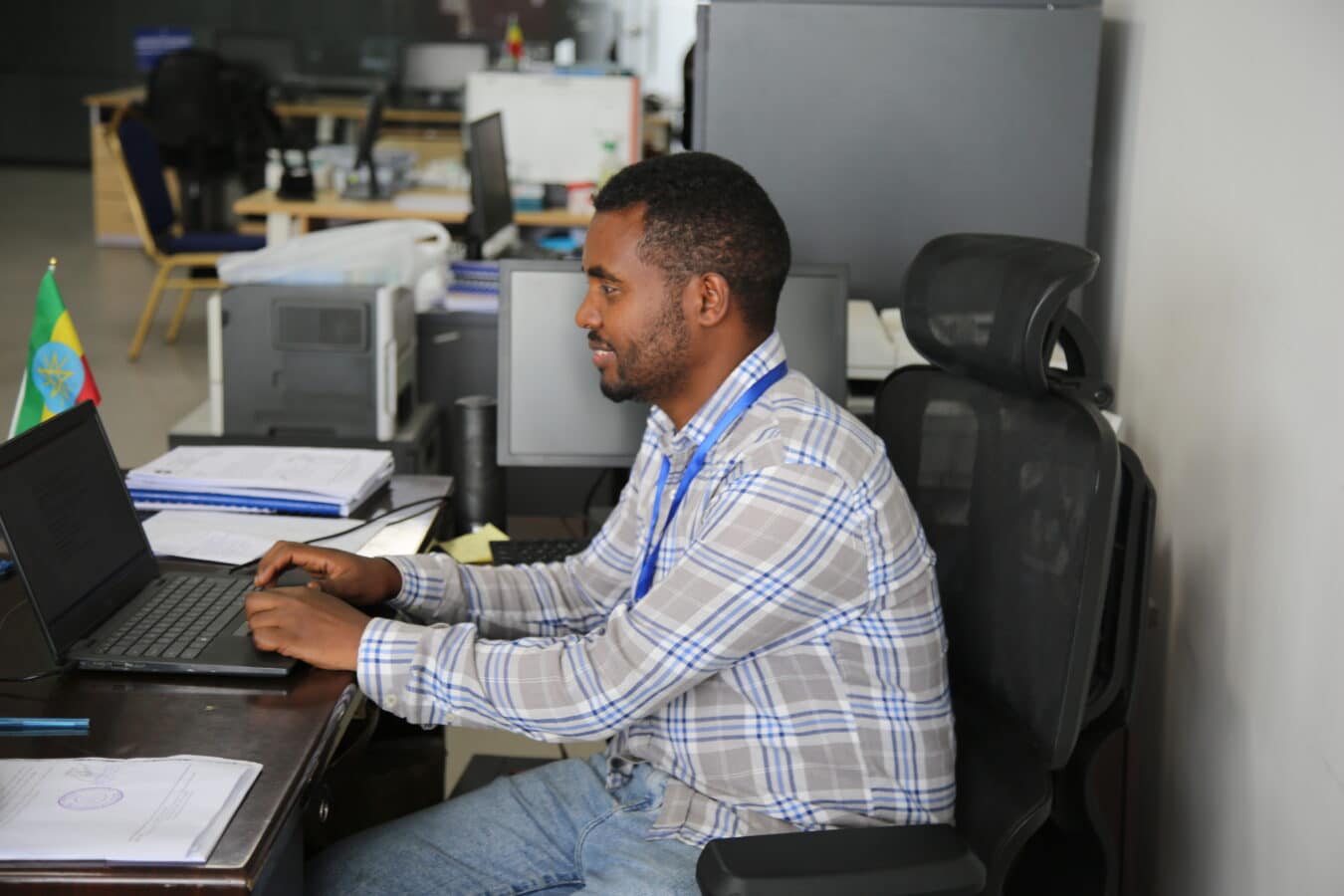 A man in a plaid shirt sits at a desk, working on a laptop in an office setting. Office equipment, documents, and stacks of papers are visible around him. An Ethiopian flag is on the left side of the desk. The background shows other workstations and office furniture.