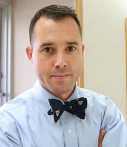 A man with short dark hair, wearing a light blue dress shirt and a dark bow tie with small shield patterns, stands indoors with his arms crossed, looking at the camera.