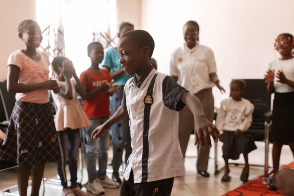 A group of children are gathered in a room, smiling and clapping. One child in the foreground is dancing, while others watch and cheer with joyful expressions. The setting appears to be indoors, with a warm and lively atmosphere.