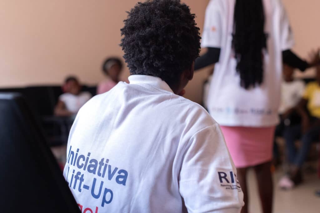 A person with curly hair, wearing a white shirt with the text "Iniciativa Lift-Up," is sitting indoors, facing a presenter in a pink skirt. Other people are sitting and listening in the background.