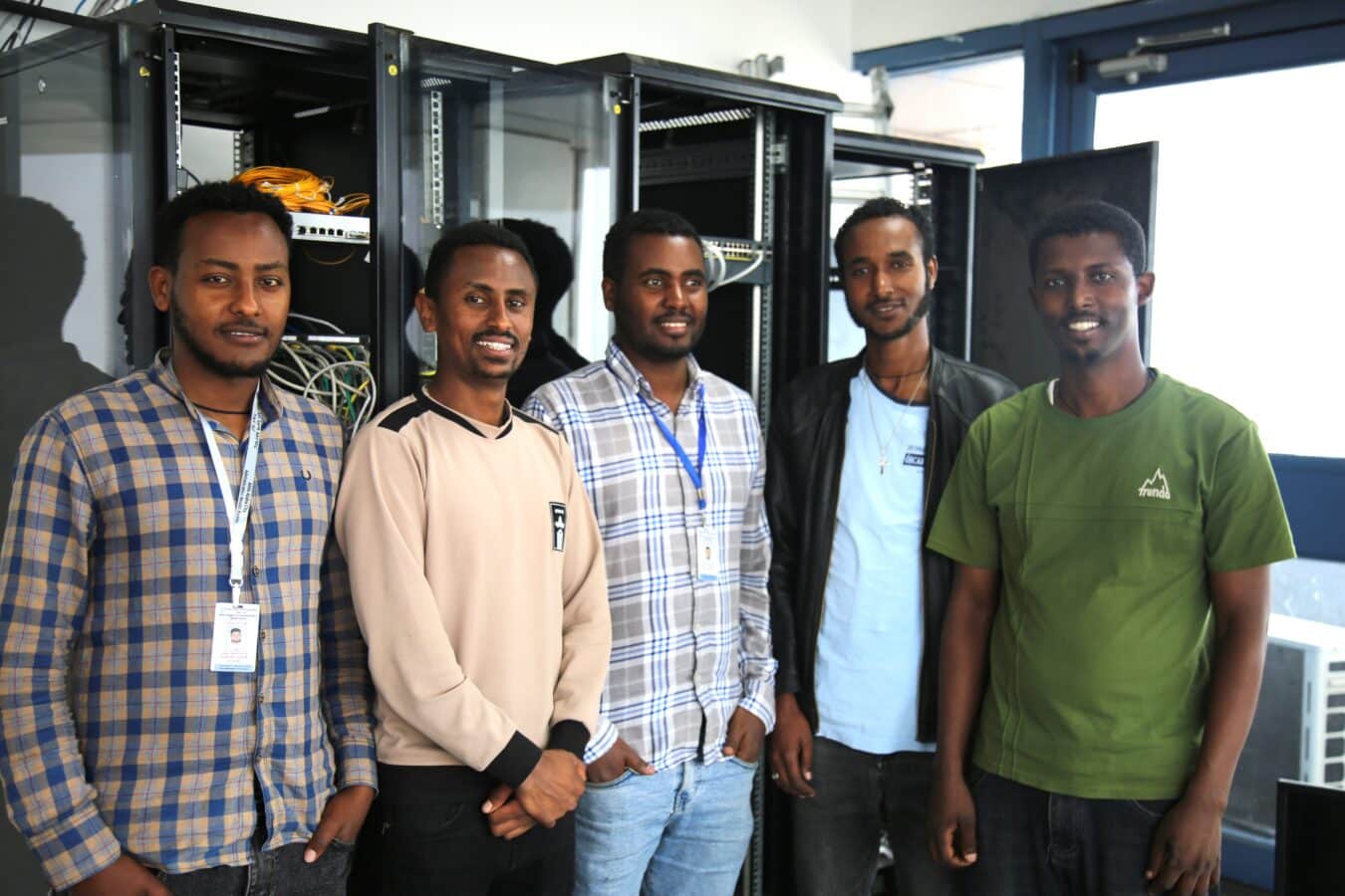 Five people stand in a room with open server racks and networking equipment. They are all smiling and appear to be relaxed. Four of them wear lanyards with ID badges. The room has a blue door, and there are various cables visible in the background.