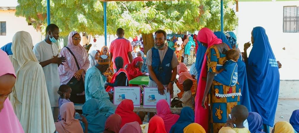 A community gathering with people in colorful attire sitting and standing under a canopy. Some are holding boxes with writing, while adults and children interact in a shaded outdoor area, surrounded by trees and buildings.