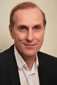 A middle-aged man with short, light brown hair is wearing a dark blazer over a white collared shirt. He is smiling slightly and looking directly at the camera against a plain, light background.