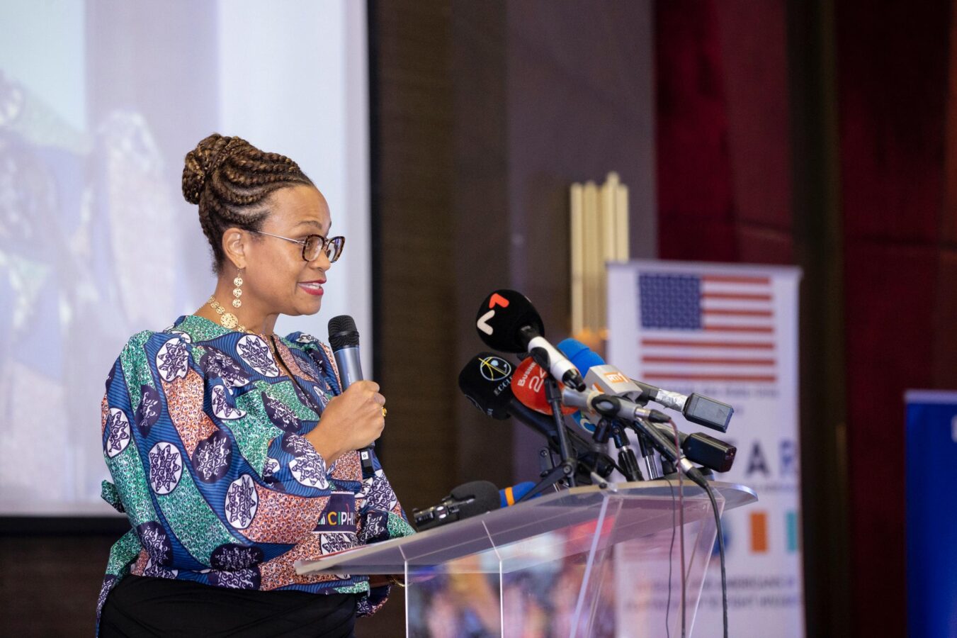 A woman with braided hair and glasses stands at a podium, speaking into a microphone. She is wearing a printed top and surrounded by several microphones. A screen and a banner with an American flag can be seen in the background.