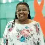 Smiling person with long hair in a floral blouse stands in front of a blurred background featuring an orange ribbon design.