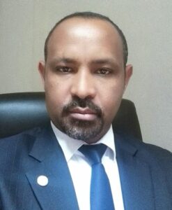 A man with a short beard wearing a suit and tie sits in an office chair facing the camera. The background is a plain wall.