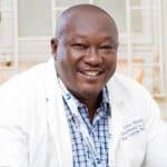 A man wearing a white lab coat and a blue checked shirt smiles at the camera, sitting indoors in front of a window with white bars.