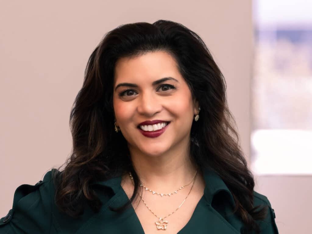 A woman with long, dark wavy hair wearing a dark green top and gold jewelry smiles at the camera. The background is softly blurred with neutral tones and a window.