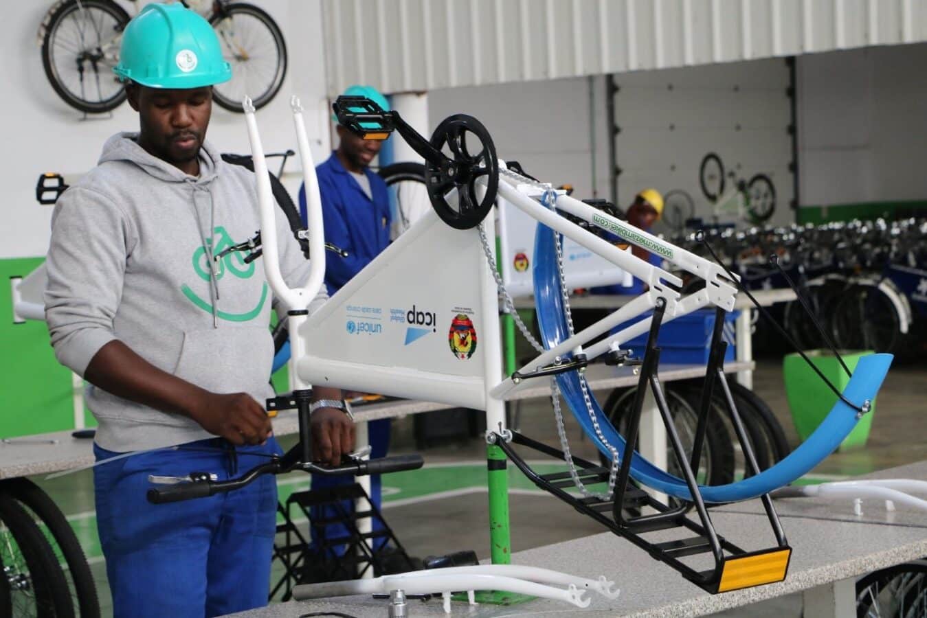 Two people in a workshop work on assembling bicycles. The person in the foreground, wearing a blue helmet and gray hoodie, adjusts the handlebars of a bike mounted on a stand. Another worker in the background is focused on a different task.