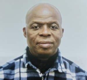 A man with a bald head and dark complexion is wearing a black and white checkered shirt. He is looking directly at the camera against a plain white background.