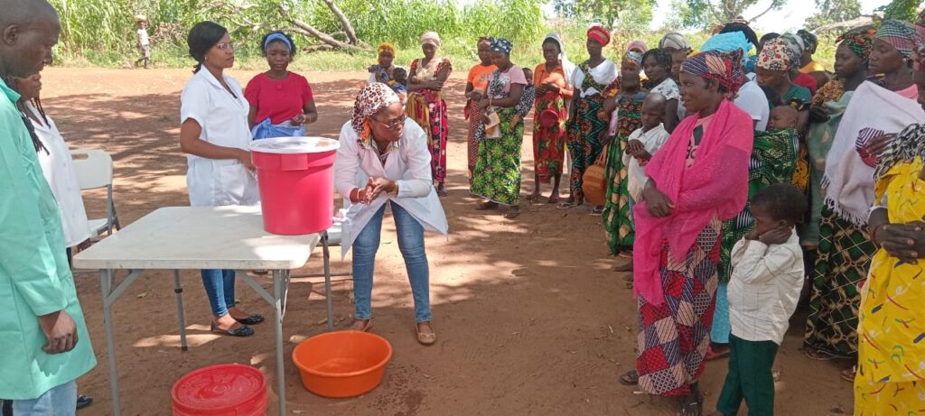 A woman in a lab coat demonstrates handwashing at a table with buckets, while a group of women and children watch attentively outdoors in a rural setting.