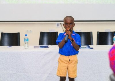 A young child stands at the front of a room holding a microphone. The child wears a blue shirt and beige shorts. Behind them is a table with water bottles, notebooks, and chairs. The background features a projector screen.