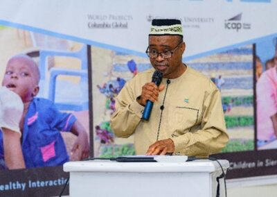 A person wearing a light beige traditional outfit and cap speaks into a microphone while standing at a podium. Behind them is a banner featuring images of community health activities and logos of various organizations, including Columbia Global and ICAP.