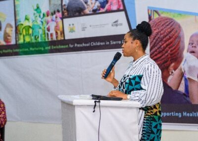 A woman in patterned clothing speaks into a microphone at a podium. Behind her, a banner displays images of programs for preschool children in Sierra Leone. Another person stands to the left, partially visible. The setting appears to be an event or conference.