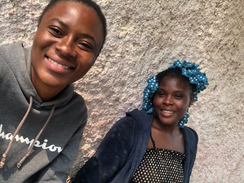 Two people smiling while standing in front of a textured wall. The person on the left is wearing a gray hoodie, and the person on the right is wearing a blue headscarf with polka dots and a dark jacket.