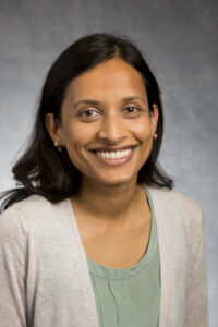 A smiling person with long dark hair, wearing a mint green top and a light gray cardigan, is shown against a plain gray background.