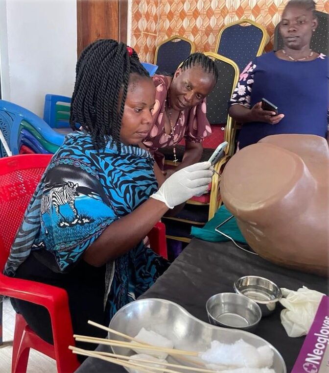 A woman wearing white latex gloves holds a smart phone up to a model of a human pelvis while other women watch her