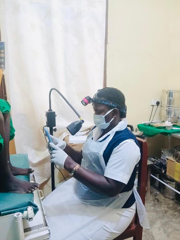 A woman wearing a mask, apron, and a light on her forehead looks at a smartphone in front of a woman standing on a table barefoot