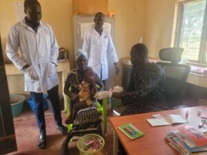 A healthcare worker hands medicine to a child sitting on a woman’s lap in a clinic, while two other medical staff in white coats stand nearby. Medical supplies and documents are on a desk in the foreground.