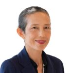 A person with short, graying hair and a warm smile is pictured against a white background. They are wearing a dark blue blazer and a black shirt.